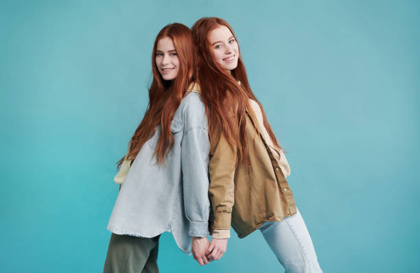Horiozntal medium long studio portrait of cheerful twin sisters wearing casual outfits standing back to back holding hands smiling at camera Twin Sisters Standing Back To Back twins back to back stock pictures, royalty-free photos & images
